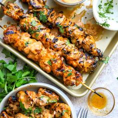 chicken skewers on a tray with dipping sauces and cilantro leaves