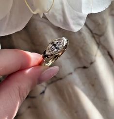 a woman's hand holding a diamond ring in front of white orchids on a marble surface