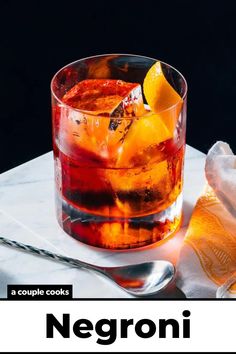 a glass filled with ice and orange slices on top of a white marble table next to a spoon