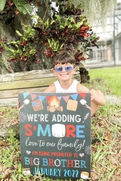 a young boy sitting in the grass holding up a sign that says we're adding smore to our family