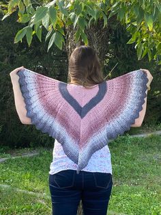 a woman standing in front of a tree with her back to the camera, holding up a pink and blue crocheted shawl