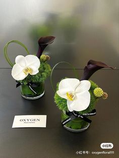 two vases with white flowers and greenery in them on a table next to a sign