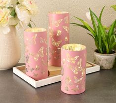 three pink vases with gold butterflies on them sitting next to a potted plant