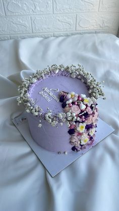 a purple cake with flowers on it sitting on top of a white sheet covered bed