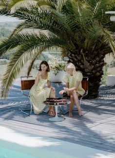 two women sitting next to each other under a palm tree on a deck near a pool