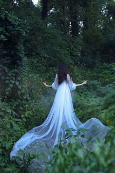 a woman in a white dress is walking through the woods with her arms spread out