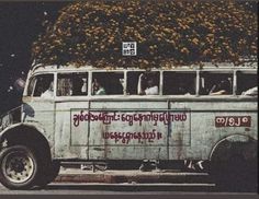 an old bus is loaded with flowers on the roof and people in the passenger seat