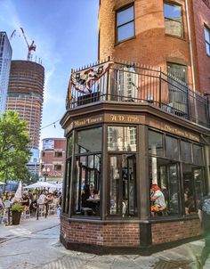 people are sitting at tables in front of a building on the street corner with tall buildings behind them