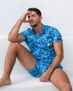 A man models the WET Stretch Shirt and matching beach shorts, sitting against a white textured wall. The vibrant blue outfit features a water-inspired pattern, designed for both comfort and fashion, perfect for resort or beachwear. Designed by DC2 Miami, a premier men's beachwear brand from Miami. Upf 50+ Short Sleeve Beachwear Tops, Upf 50+ Short Sleeve Beach Tops, Casual Short Sleeve Swimwear With Upf 50+, Casual Blue Short Sleeve Swimwear, Cotton Tops With Upf 50+ For Summer, Gym Tank Tops, Tailored Shorts, Men's Button Down Shirt, Swim Brief