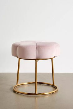 a pink ottoman with gold legs and a round foot rest on a concrete floor in front of a white wall