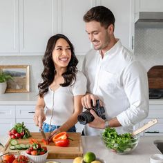 a man and woman standing in the kitchen preparing food on a cutting board while holding a camera