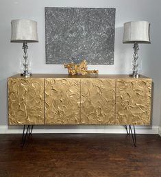 a gold sideboard with two lamps on it in front of a painting and lamp shades