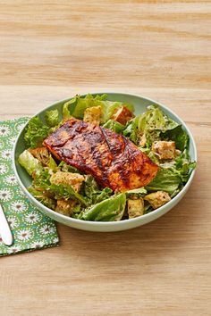 a salad with meat and lettuce in a bowl next to a fork on a wooden table