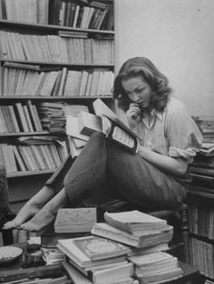 a woman sitting on the floor reading a book and talking on her cell phone in front of a bookshelf