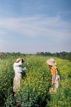 two women are standing in a field with yellow flowers and one is holding a camera