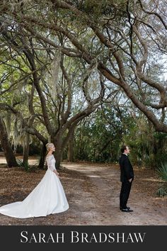 a bride and groom standing under the trees in their wedding gowns, looking into each other's eyes