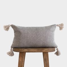 a grey and white pillow with tassels sitting on top of a wooden stool