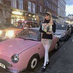 a woman standing next to a pink car on the side of a road in front of parked cars