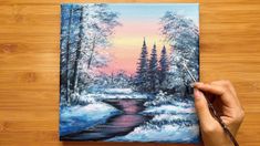 someone is painting a winter scene with water and trees in the background on a wooden surface