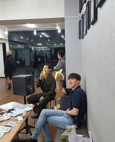 two men sitting at a table with bananas in their hands and people standing around them