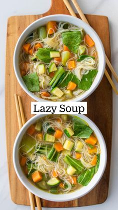 two bowls of vegetable noodle soup on a cutting board with chopsticks next to it