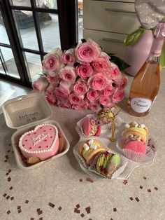 a table topped with pink roses and cupcakes next to a bottle of wine