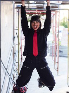 a woman in black jacket and red tie standing on snowboard with hands up above her head