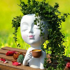 a white statue holding a cup with plants growing out of it's head on top of a wooden table