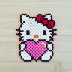 a cross stitch hello kitty with a heart on it's chest sitting on a wooden surface