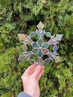 a hand holding a stained glass snowflake in front of a green tree with lots of leaves