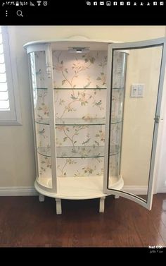 a white corner cabinet with glass doors and flowers on the wall next to a wooden floor