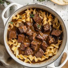 a pot filled with beef and noodles on top of a wooden table next to bread