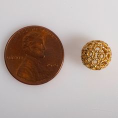 a penny sitting next to a small gold bead on a white surface with a coin in the background