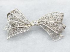 Add a touch of elegance and whimsy to any outfit with this bow brooch. The delicate filigree detailing and sparkling European Cut diamond make this brooch a timeless statement piece. Perfect for any occasion! Metal: 14K White Gold Gem: European Cut Diamond .10 Carats, SI1 in Clarity, I in Color Gem Measurements: 3.2 mm, Round Measurements: 44 x 23 mm Marks: "X14K" Stamped on the pin guard SKU #: A39573 Each piece has been identified and graded by a Graduate Gemologist who has been certified by the Gemological Institute of America (GIA). We have six brick-and-mortar storefronts in Maine, Massachusetts, and New Hampshire and have been in business for over 25 years! Please visit our Shop's About Page or our website for more information about our jewelry. For questions about diamond grading, w Classic Wedding Brooches With Intricate Design, Elegant White Gold Brooch With Intricate Design, White Gold Brooches With Intricate Design For Wedding, Elegant White Gold Brooches With Intricate Design, Wedding Brooch With Intricate Diamond Design, White Gold Wedding Brooches With Intricate Design, Elegant Brooches With Intricate Design For Anniversary, Elegant Filigree Brooches For Evening, Wedding Diamond Brooch With Intricate Design