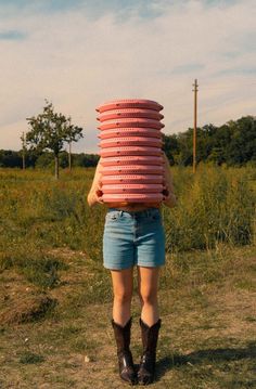 a woman carrying a stack of pink donuts on her head in the middle of a field