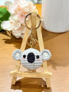 a crocheted koala keychain hanging from a wooden stand with flowers in the background