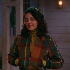 a woman standing in front of a house wearing a green hat and plaid jacket with her hands on her hips