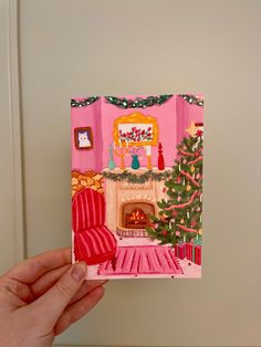 a hand holding up a christmas card with a fireplace in the corner and pink walls