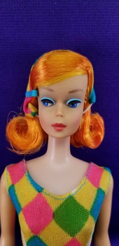 a doll with orange hair and blue eyes wearing a multicolored dress on a purple background