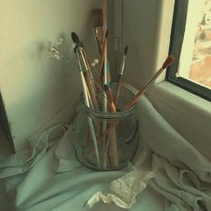 a glass vase filled with lots of brushes on top of a cloth covered table next to a window