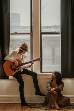 a woman sitting on the floor next to a man playing an acoustic guitar in front of a window