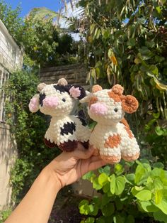 a hand holding two small stuffed animals in front of some bushes and trees with green leaves