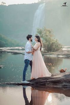 a man and woman standing next to each other near a body of water with a waterfall in the background