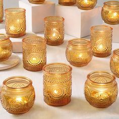 there are many gold glass candles on the table with white boxes around them and one candle is lit