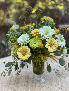 a glass vase filled with yellow and white flowers on top of a wooden table covered in greenery