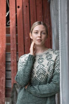 a woman standing in front of a red fence wearing a green sweater and holding her hand to her ear