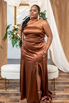 a woman standing in front of a white chair wearing a brown dress and gold earrings