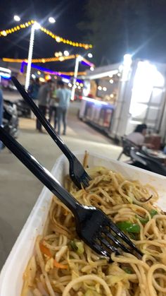 a fork and some noodles in a white container on the street with people walking by