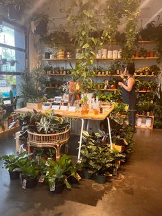 a woman standing in front of a table filled with lots of potted plants and greenery