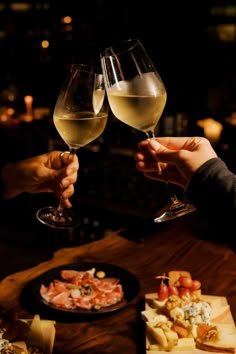 two people toasting wine glasses over cheese and crackers on a table in a dimly lit room